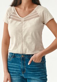 Top beige de manga corta con escote en V, que presenta detalles de encaje y un corte recto. Combinado con jeans de mezclilla azules.