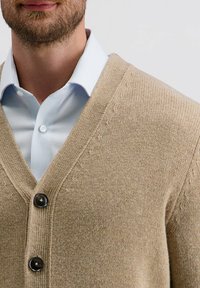 Beige gebreide cardigan met een V-hals, met een gestructureerd oppervlak en twee zwarte knopen. Gelaagd over een lichtblauw gestreept shirt.