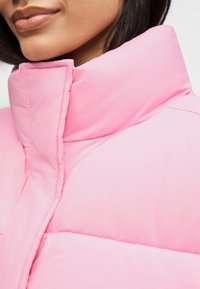 Κοντινό πλάνο ενός ατόμου που φοράει μπουφάν τύπου πάπλωμα με υψηλό γιακά, σε ανοιχτό ροζ χρώμα, παρουσιάζοντας την υφή και το σχέδιο του γιακά του μπουφάν.