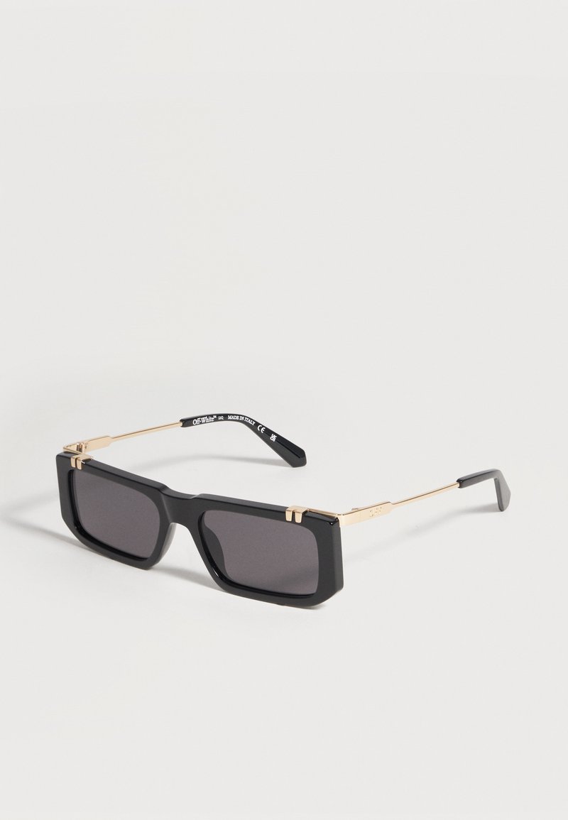 Lunettes de soleil rectangulaires noires avec montures épaisses et branches fines dorées, présentant des détails de marque sur les branches.