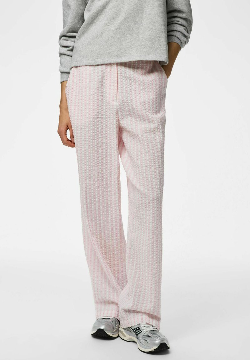 Personne portant un pantalon large texturé rose clair, un sweat-shirt gris et des baskets gris argenté, debout avec une main dans la poche.