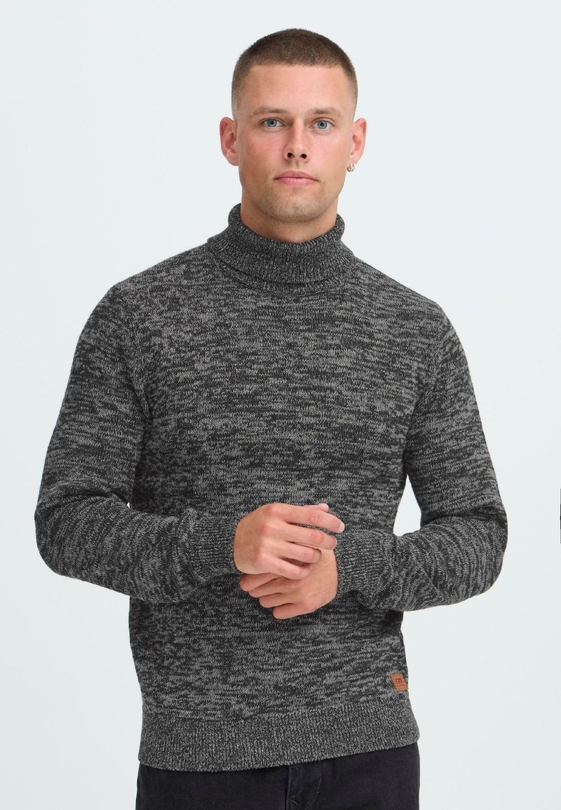 Jeune homme aux cheveux courts portant un pull col roulé gris chiné, debout les mains jointes contre un fond blanc uni.