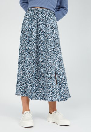 Jupe mi-longue bleue à fleurs avec une fente latérale, associée à des baskets blanches et un pull en maille bleu, portée par une personne debout.