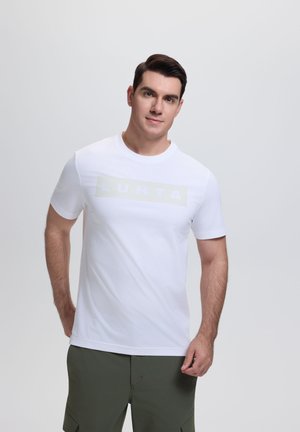 Mann mit kurzen dunklen Haaren, der ein weißes Luhta-T-Shirt und olivgrüne Hosen trägt und vor einem schlichten hellen Hintergrund steht.