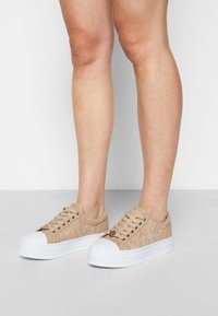 Béžové krajkové tenisky s bílými gumovými podešvemi, které mají strukturovaný vzor a kovový akcent na boku, stylizované s kulatými špičkami a tkaničkami.