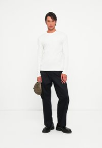 Jonge man die tegen een witte achtergrond staat, gekleed in een witte lange mouw shirt, zwarte broek, zwarte schoenen en een bruine geruite pet vasthoudt.