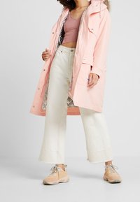 Jasnoróżowa wodoodporna parka z kapturem z sztucznego futra, srebrną podszewką, przednimi kieszeniami, połączona z kremowymi spodniami typu wide-leg i brązowymi sneakersami.