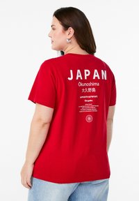 Sarkanais kokvilnas T-krekls ar uzrakstu "JAPAN" baltajās burtiem aizmugurē, kā arī tekstu un grafiskajiem elementiem par Ōkunoshimu. Brīva piegriezuma, apaļa kakla izgriezums.