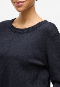 Dark navy sweater made of textured knit fabric, featuring a ribbed round neckline and subtle detailing near the shoulder seam.
