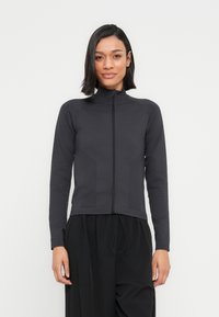 Femme aux cheveux noirs raides portant une veste noire à manches longues zippée et un pantalon noir ample, debout devant un fond blanc uni.