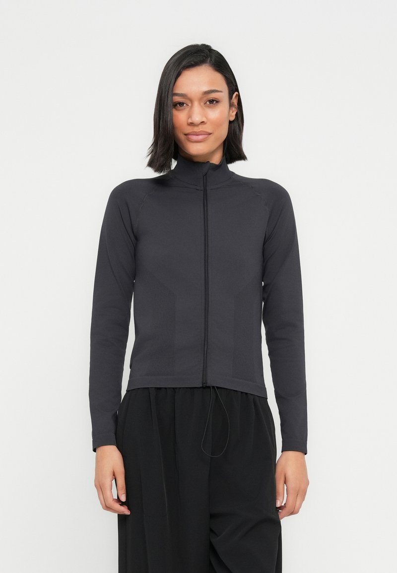 Femme aux cheveux noirs raides portant une veste noire à manches longues zippée et un pantalon noir ample, debout devant un fond blanc uni.