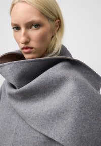 Grå ullkappa med hög krage, med en mjuk textur och en draperad design. Insidan har en kontrasterande ljusare nyans.