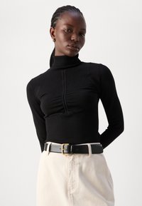 Zwarte geribde coltrui met textuurdetails, gecombineerd met een zwarte riem en lichtbeige hoge taillebroek.