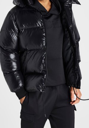 Hombre con chaqueta acolchada negra brillante sobre una sudadera con capucha negra y pantalones cargo negros, con una mano en el bolsillo y la otra sujetando un cordón.