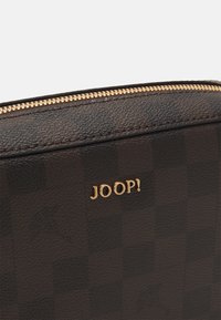 Matériau à motif écossais brun avec quincaillerie dorée. Présente un logo "JOOP!" bien en évidence, mettant en valeur la fermeture éclair et la finition texturée.