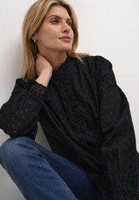 Juoda palaidinė su siuvinėtų skylučių raštu, aukštu kaklu ir mygtukų uždarymu. Su pūstomis rankovėmis su elastinėmis apykaklėmis. Dėvima su mėlynais džinsais.