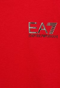 Červené bavlněné tričko s výrobním logo ve stříbrné barvě obsahující text "EA7" a "EMPORIO ARMANI". Hladká textura, přiléhavý design, bez vzorů či dalších akcentů.