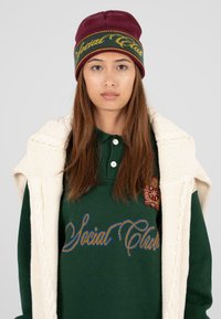 Bordeaux gebreide beanie met groene band en gele tekst, groene polo met blauwe borduursel, en crèmekleurige gebreide cardigan over de schouders.