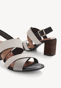 Paire de sandales ouvertes pour femme avec des lanières croisées beige et noires, une boucle ajustable et un talon épais marron sur fond blanc.