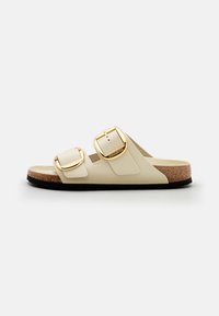 Twee-kleurige sliders in crèmewit en beige met gouden gespen, een kurken voetbed en een textuur van zwart rubberen zool.