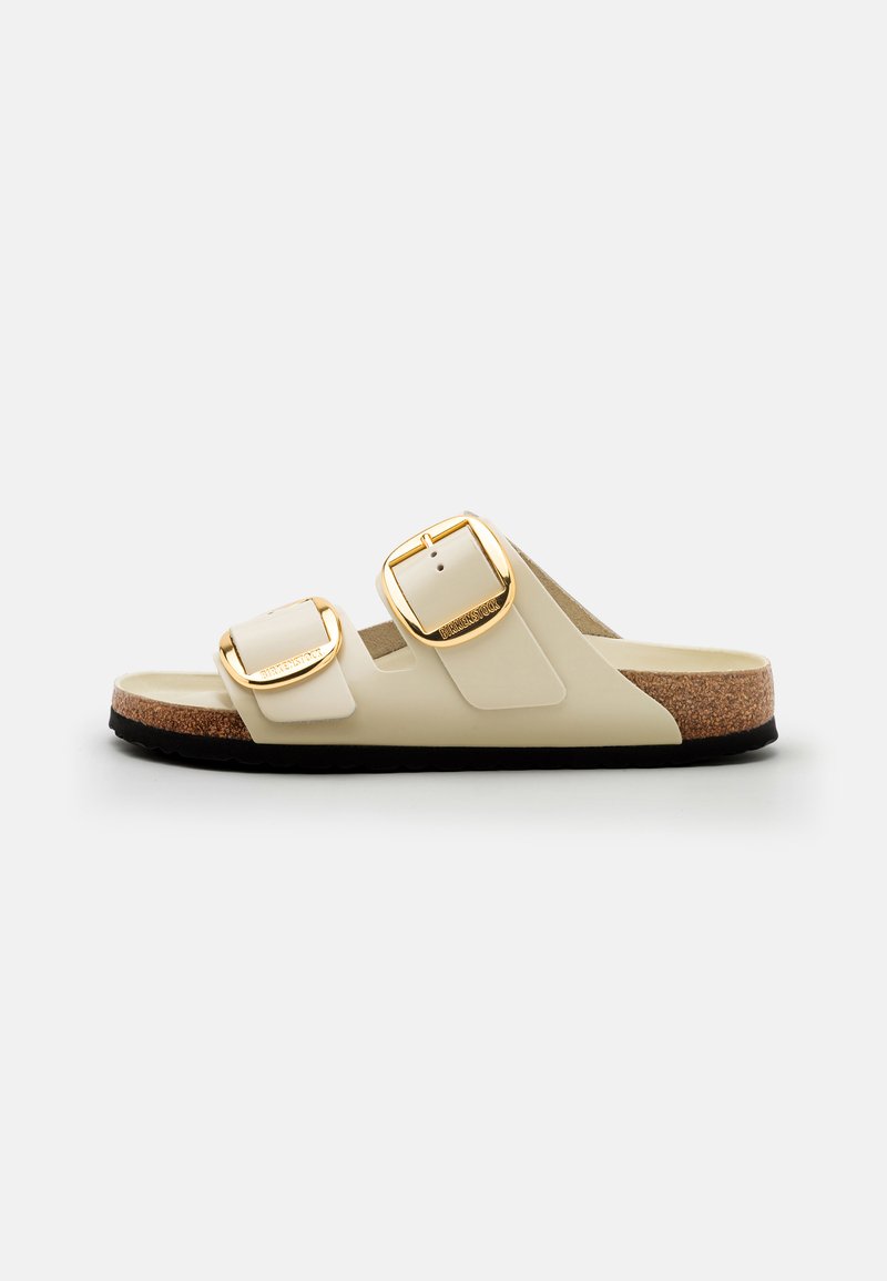 Twee-kleurige sliders in crèmewit en beige met gouden gespen, een kurken voetbed en een textuur van zwart rubberen zool.