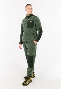 Hombre de pie con chaqueta de forro polar verde y pantalones con parches negros, mirando de lado, y botas de senderismo amarillas.