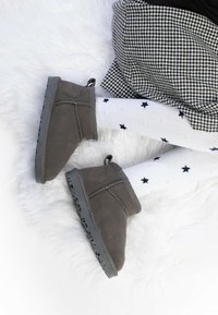 Botas de tobillo de ante gris con suela texturada, complementadas con calcetines blancos con estrellas marinas. El fondo es una suave textura de piel blanca.