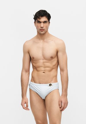 Izmos férfi sötét hajjal, fehér úszónadrágot visel, amelyen egy kis díszítő embléma van a bal csípőn, egyszerű háttér előtt áll.