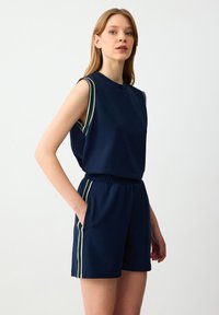 Top senza maniche blu navy con strisce verdi e bianche sulle spalle, abbinato a pantaloni corti coordinati con tasche laterali e dettagli a strisce.