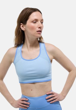 Femme portant un soutien-gorge de sport bleu clair et un legging, posant les mains sur les hanches et regardant sur le côté, devant un fond clair uni.