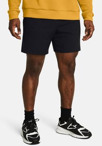 Schwarze Sportshorts mit flacher Vorderseite, ohne Taschen, getragen mit schwarz-weißen Sneakers und schwarzen Socken. Der Pullover ist senfgelb.