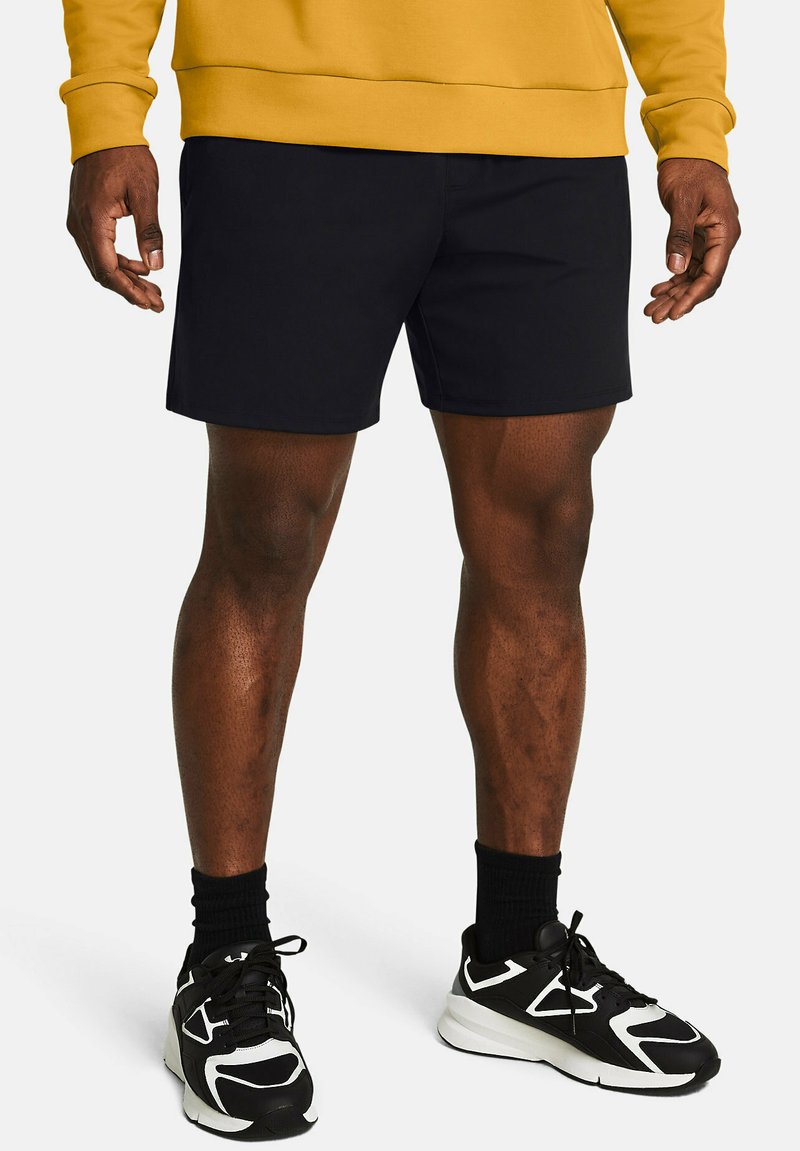 Schwarze Sportshorts mit flacher Vorderseite, ohne Taschen, getragen mit schwarz-weißen Sneakers und schwarzen Socken. Der Pullover ist senfgelb.