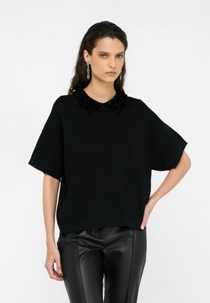 Schwarzes oversize Oberteil mit einem strukturierten schwarzen Kragen. Das Model trägt schwarze Kunstlederhosen. Einfaches Design, kurze Ärmel, glänzender Stoff.