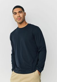 Navy long-sleeve t-shirt made from a smooth fabric with a crew neckline and a subtle logo on the chest. Paired with beige pants.