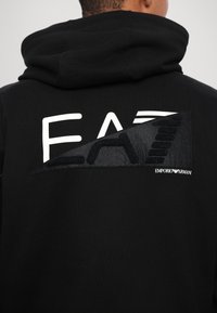 Personne portant un sweat à capuche noir avec un grand logo EA7 Emporio Armani blanc et noir dans le dos.