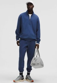 Felpa blu in pile con zip e pantaloni joggers coordinati con polsini. Borsa da palestra grigia e sneakers grigie con accenti arancioni completano l'outfit.