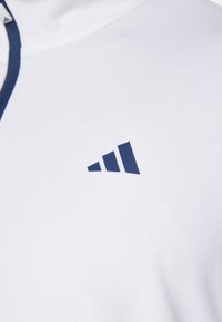 Biele športové látky s textúrovaným povrchom, s námorníckou modrou trojuholníkovou logom na ľavej strane a kontrastným námorníckym zipsom.