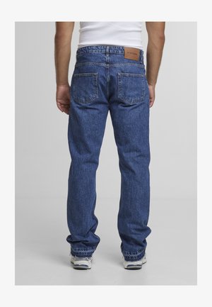 Man wearing loose-fit blue jeans with back pockets, standing, visible from waist to shoes against plain background.