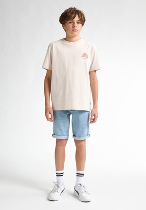 T-shirt di cotone rosa chiaro con una piccola grafica sul lato sinistro del petto, abbinata a pantaloni corti in denim arrotolati, sneakers bianche e calzini a righe.