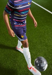 Blauwe en gestreepte korte-sleeve voetbalshirt, witte shorts, witte sokken en rode voetbalschoenen; speler dribbelt een zwart-wit voetballetje op kunstgras.