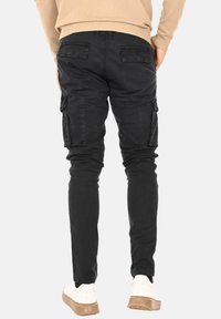 Pantaloni cargo neri in cotone, caratterizzati da un design slim-fit, tasche laterali e una finitura leggermente testurizzata. Indossati con scarpe bianche.