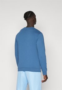 Sudadera azul con mangas largas, escote redondo y puños acanalados. La tela parece suave y lisa. Se lleva con pantalones azul claro.