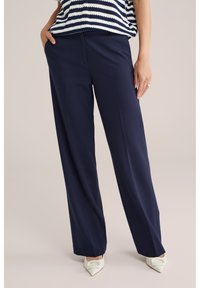 Femeie purtând pantaloni largi bleumarin, top cu dungi bleumarin și alb, și pantofi cu toc alb cu vârf ascuțit, stând cu o mână în buzunar.