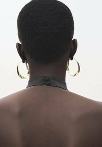 Čierny kožený halter top s detailom gombíka na chrbte, skombinovaný s veľkými zlatými kruhovými náušnicami. Hladká textúra pokožky viditeľná na krku.