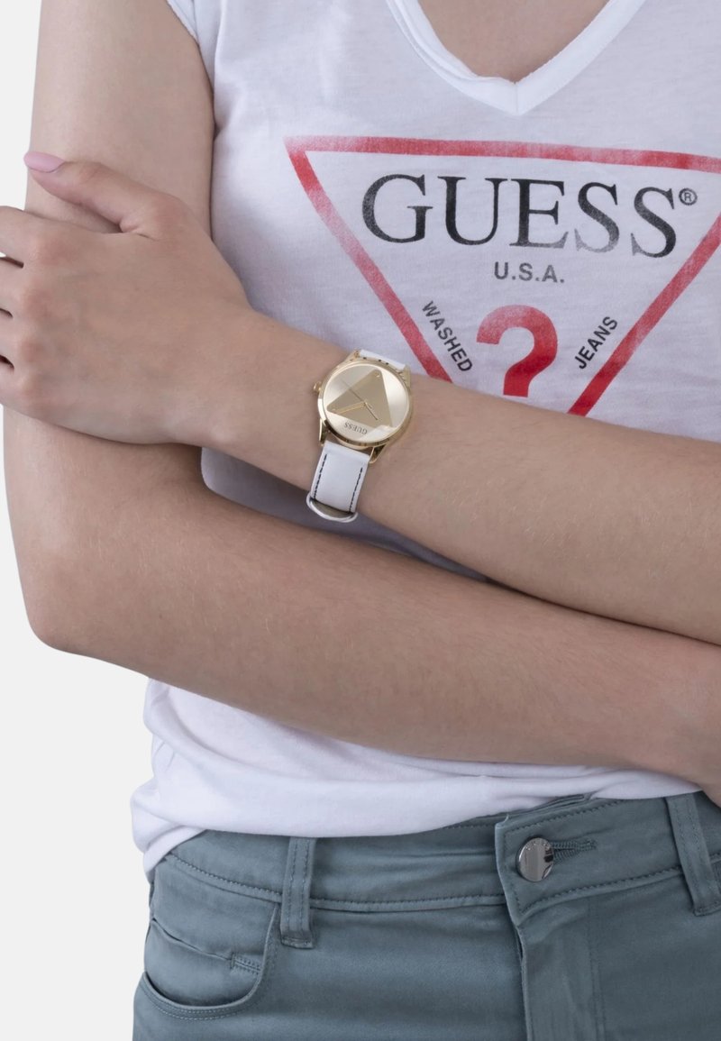 Goudkleurig kwarts horloge met driehoekige wijzerplaat en witte leren band. De pols is gedeeltelijk verborgen door een t-shirt met een driehoekig logo.