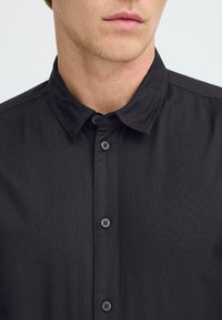 Black collared shirt with a textured fabric, featuring a row of black buttons down the front, and visible seams at the collar and shoulders.