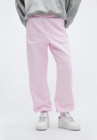 Άτομο που φορ άει ροζ ανοιχτό χρώμα φόρμας με ελαστικά τελειώματα, λευκά αθλητικά παπούτσια και γκρι φούτερ με κουκούλα μπροστά σε λευκό απλό φόντο.