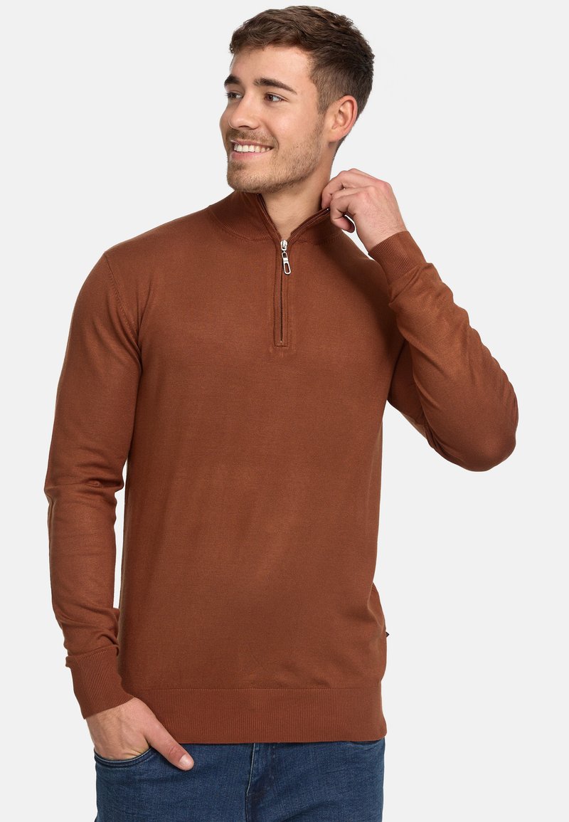 Brauner Pullover mit hohem Kragen und halbem Reißverschluss. Verfügt über eine gerippte Textur und taillierte Ärmel, kombiniert mit blauen Jeans.