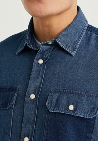 Mörkblå denimskjorta med klassisk krage, knäppning framtill och två bröstfickor med ljusa knappar och viss texturvariation.