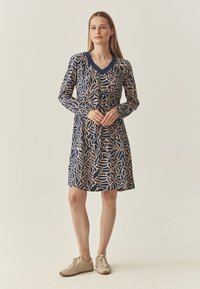 Marineblaues Kleid mit cremefarbenem botanischem Muster, V-Ausschnitt, langen Ärmeln und gerafftem Bund. Tragen mit hellbeigen Schuhen. Schlichte und lockere Passform.